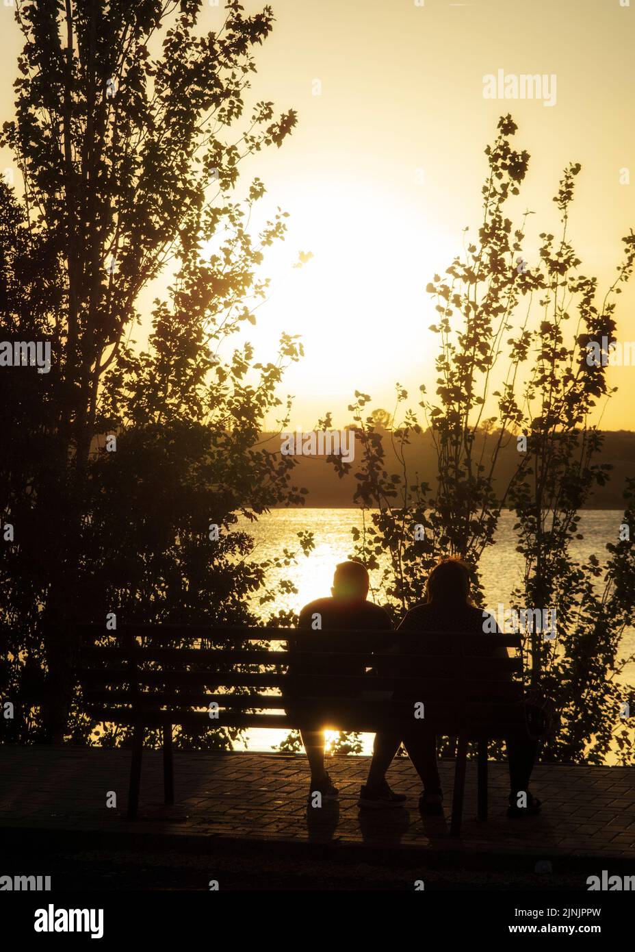 Silhouettes of a couple sitting on a bench enjoying the sunset at the lake in Arcos de la Frontera. Stock Photo