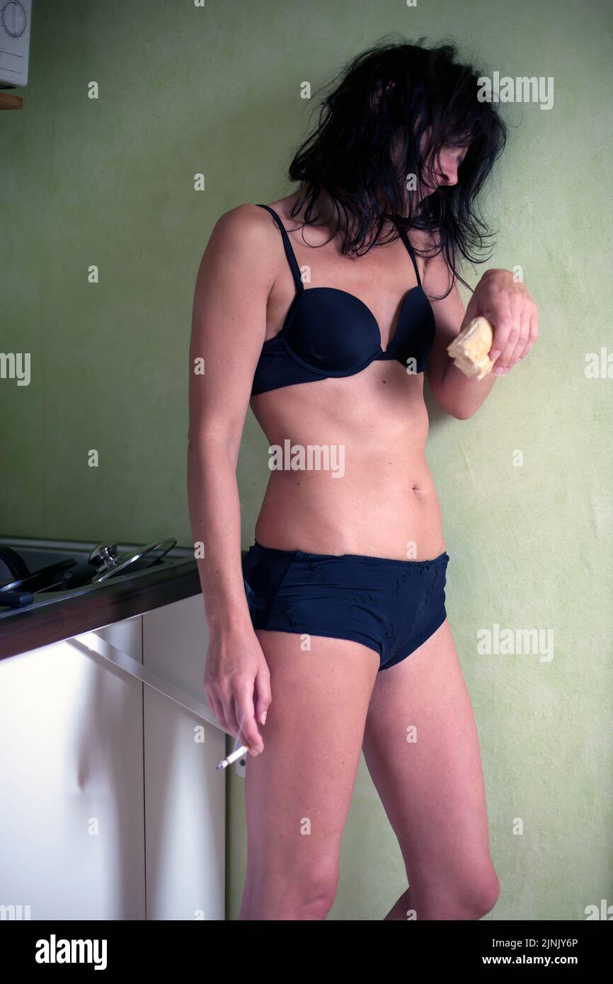 young woman standing in kitchen looking tired smoking cigarette and eating bread Stock Photo