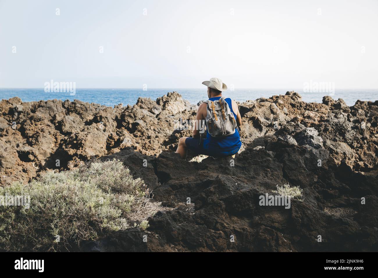 Hiker with hat is sitting on volcanic rock, has backpack on his back ...