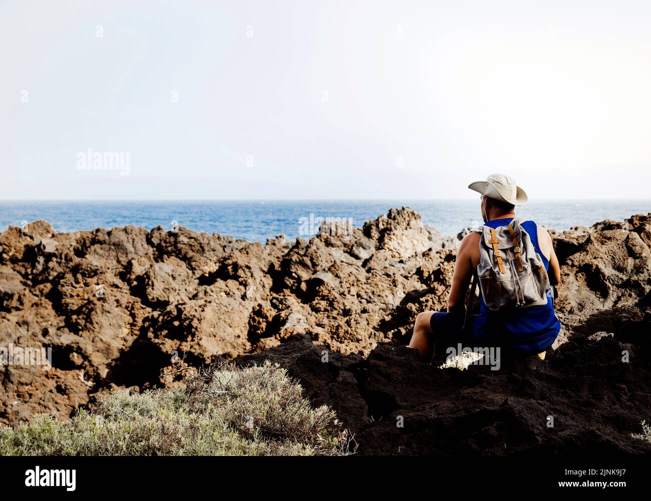 Hiker with hat is sitting on volcanic rock, has backpack on his back ...