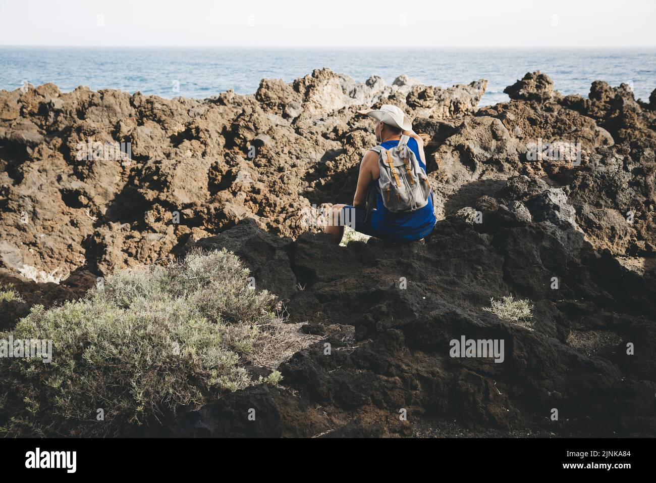 Hiker with hat is sitting on volcanic rock, has backpack on his back ...