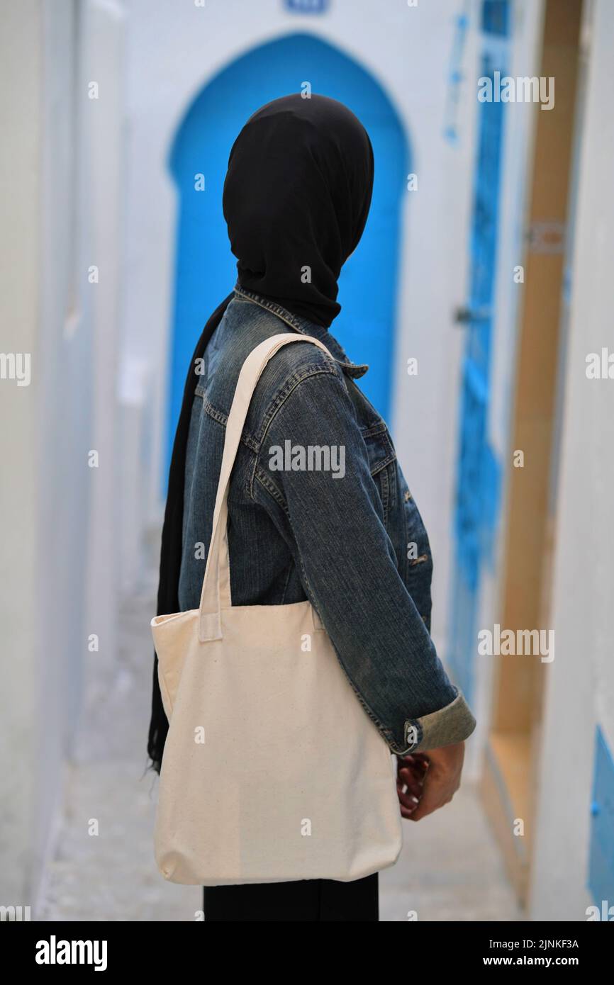 A Moroccan girl wearing a hijab carrying a canvas bag and looking at a blue Moroccan door Stock Photo