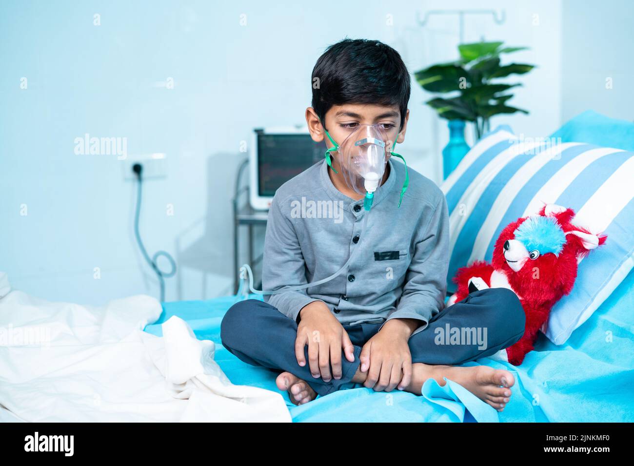 Worried sick kid with oxygen mask sitting on bed at hospital ward - concept of breathing problems, respiratory infection and health care treatment. Stock Photo