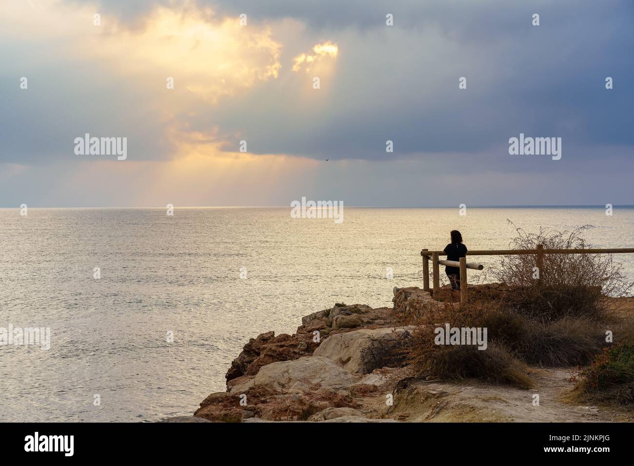 Woman looking to the horizon from a viewpoint at golden sunset over the sea. Stock Photo