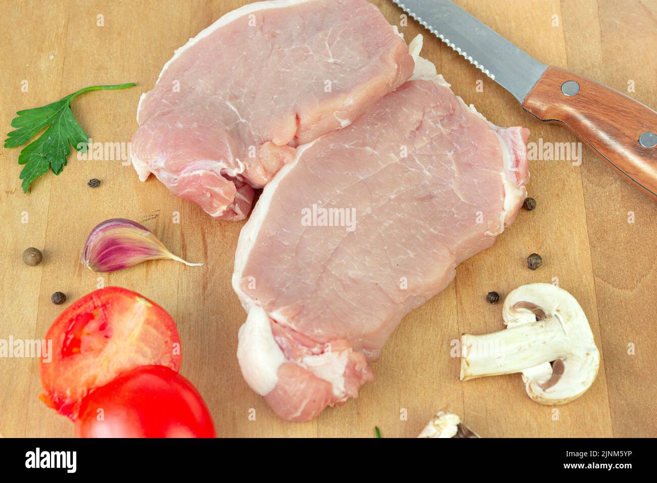 Raw pork chops of meat with garlic, tomato, spice, pepper, champignon on round cutting board with meat knife Stock Photo