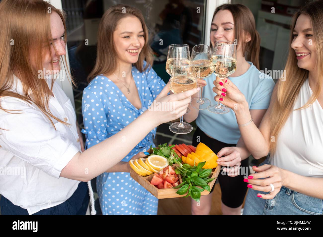 Portrait of young joyful amazing women holding vegetables, clinking glasses of white wine, standing near house. Party. Stock Photo