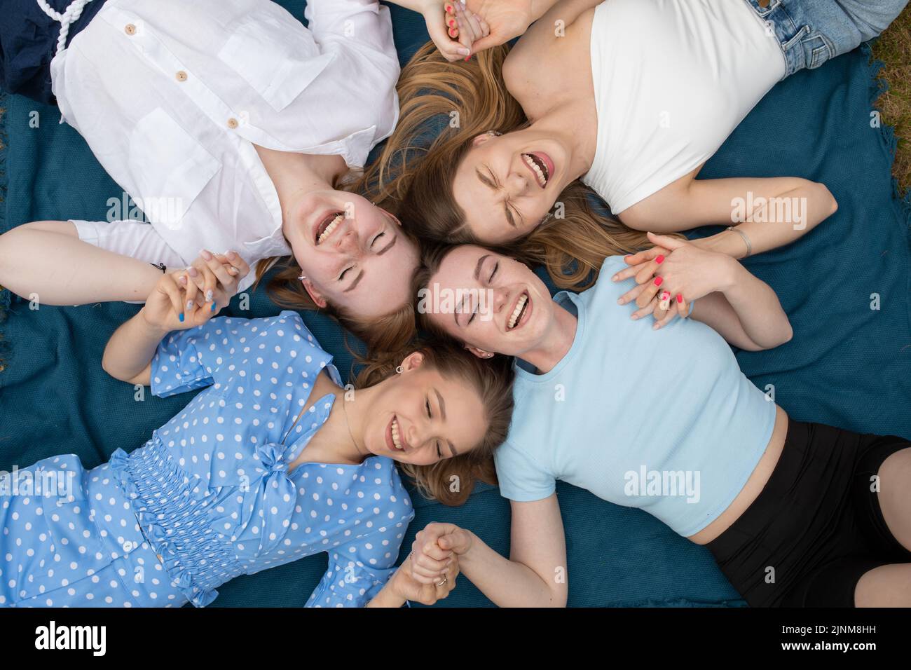 Top view of young laughing stunning women holding hands together, lying in circle on blue blanket with closed eyes. Stock Photo