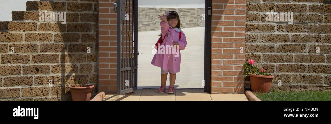 Image of an adorable little girl in a pink apron and backpack who greets her parents on her first day of school. Horizontal banner Stock Photo