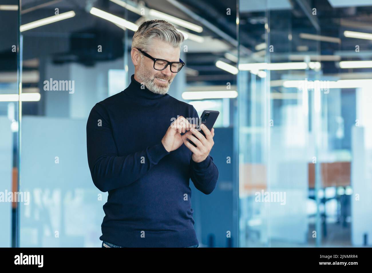 Senior gray-haired businessman in office using phone, happy investor reading message and smiling, broker at work looking at window in glasses Stock Photo