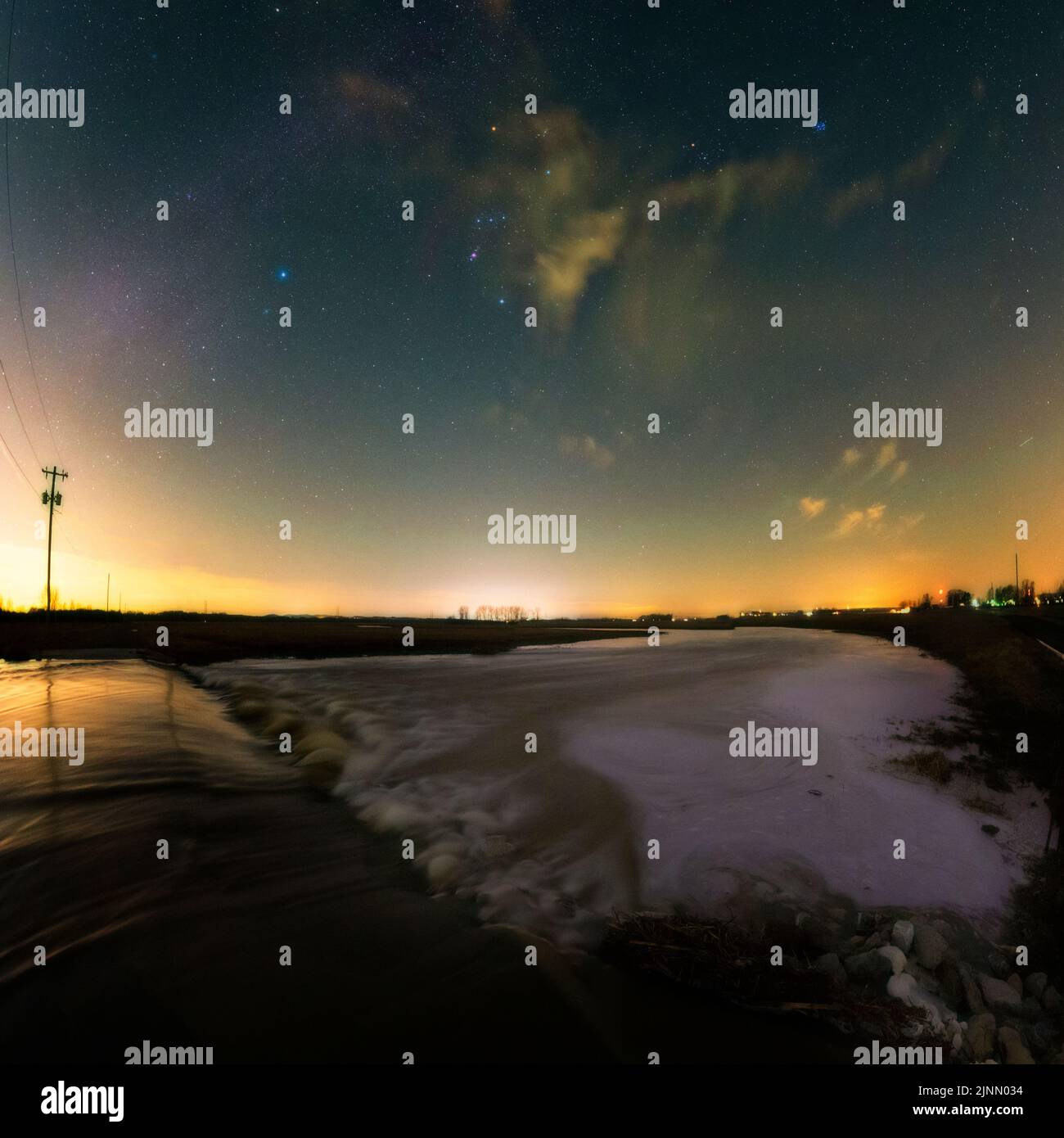 The stars of winter preside over a field that is filling with flood water.  Clouds help ground the scene.  The water looks glassy. Stock Photo