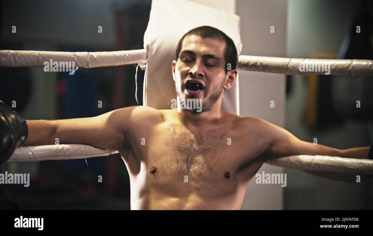A man boxer sitting on the ring in the corner with a teeth guard in the