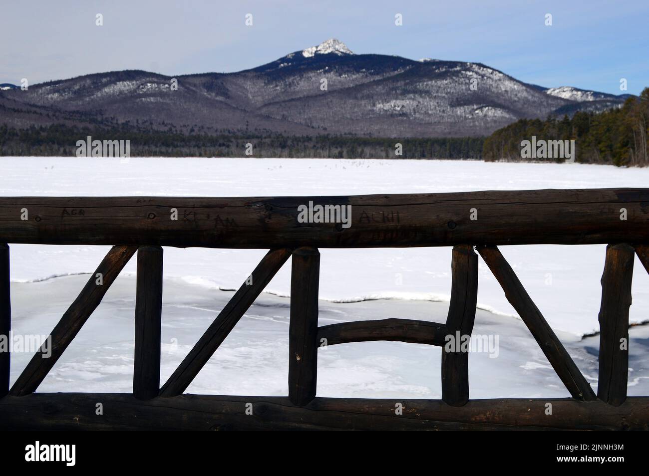 Wooden logs form a railing on a bridge that spans a small tributary stream of a frozen lake at the base of a mountain in New England Stock Photo