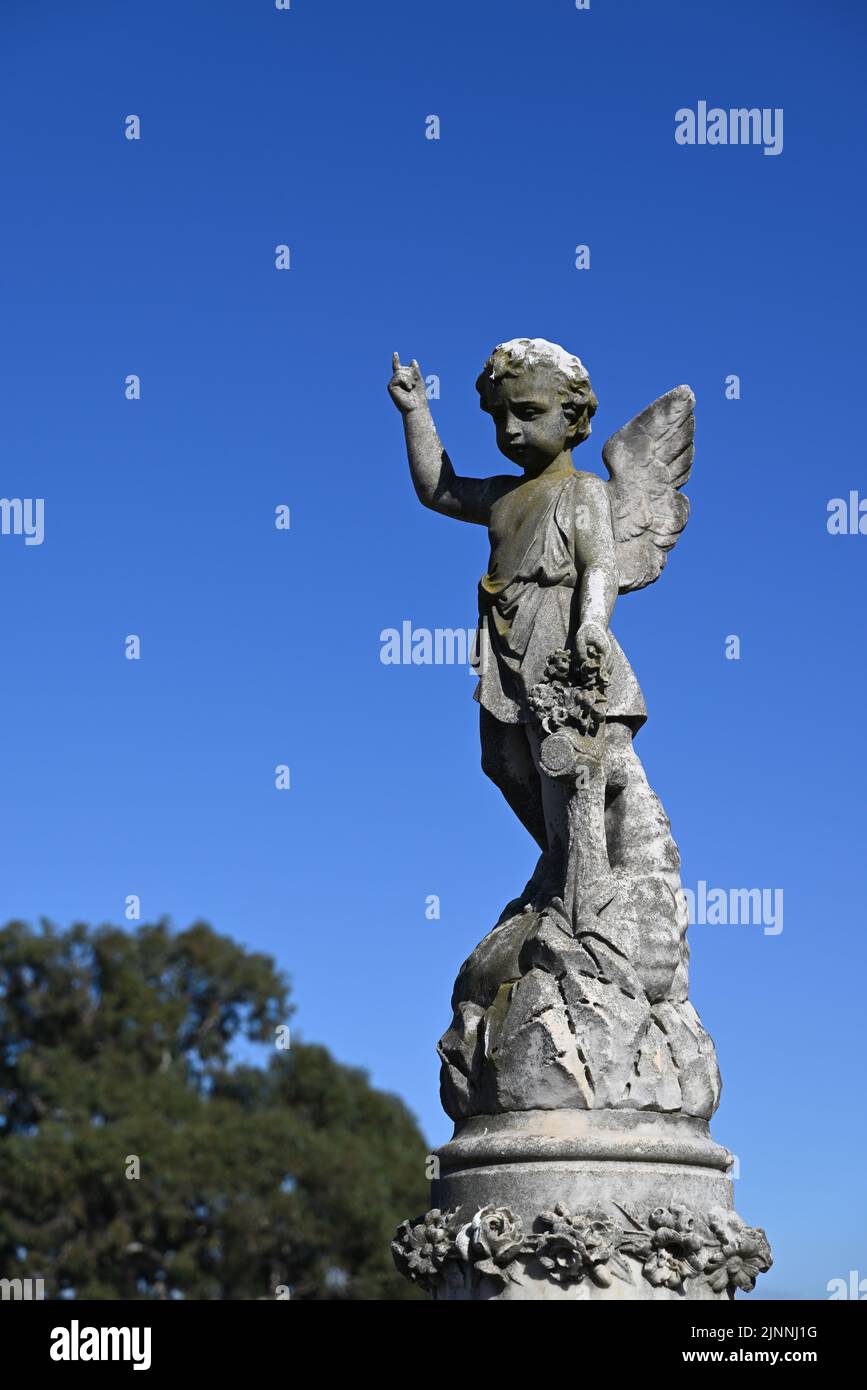 Stone sculpture of a child angel, or cherub, pointing skyward whilst looking down, atop a grave during a clear day Stock Photo
