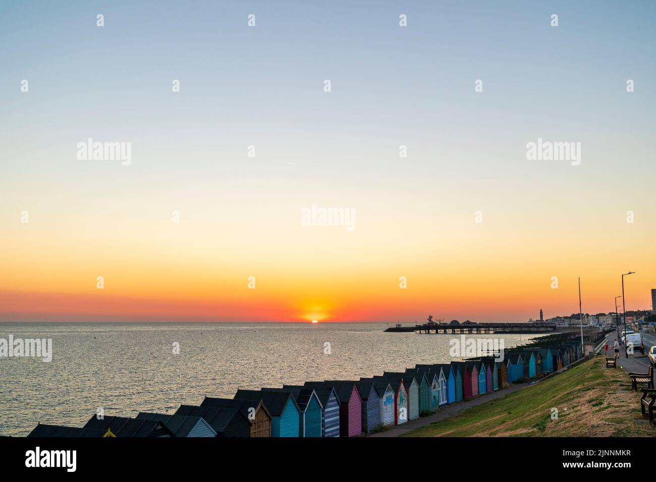 Sunrise over the North sea at Herne Bay in Kent. Sun rising above the sea with the silhouette of the Herne Bay pier and harbour jetty. Sky is orange turning gradually into blue. A row of multi-coloured beach huts in the foreground. Stock Photo