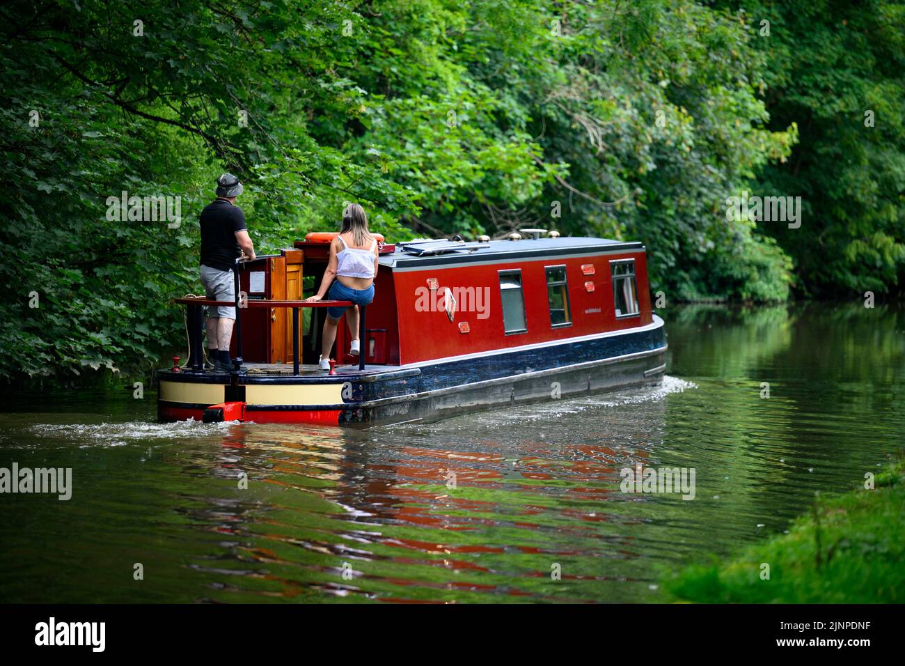 Couple relaxing on red cruiser-style narrow boat moving slowly along scenic quiet rural Leeds Liverpool Canal - Bingley, West Yorkshire, England, UK. Stock Photo