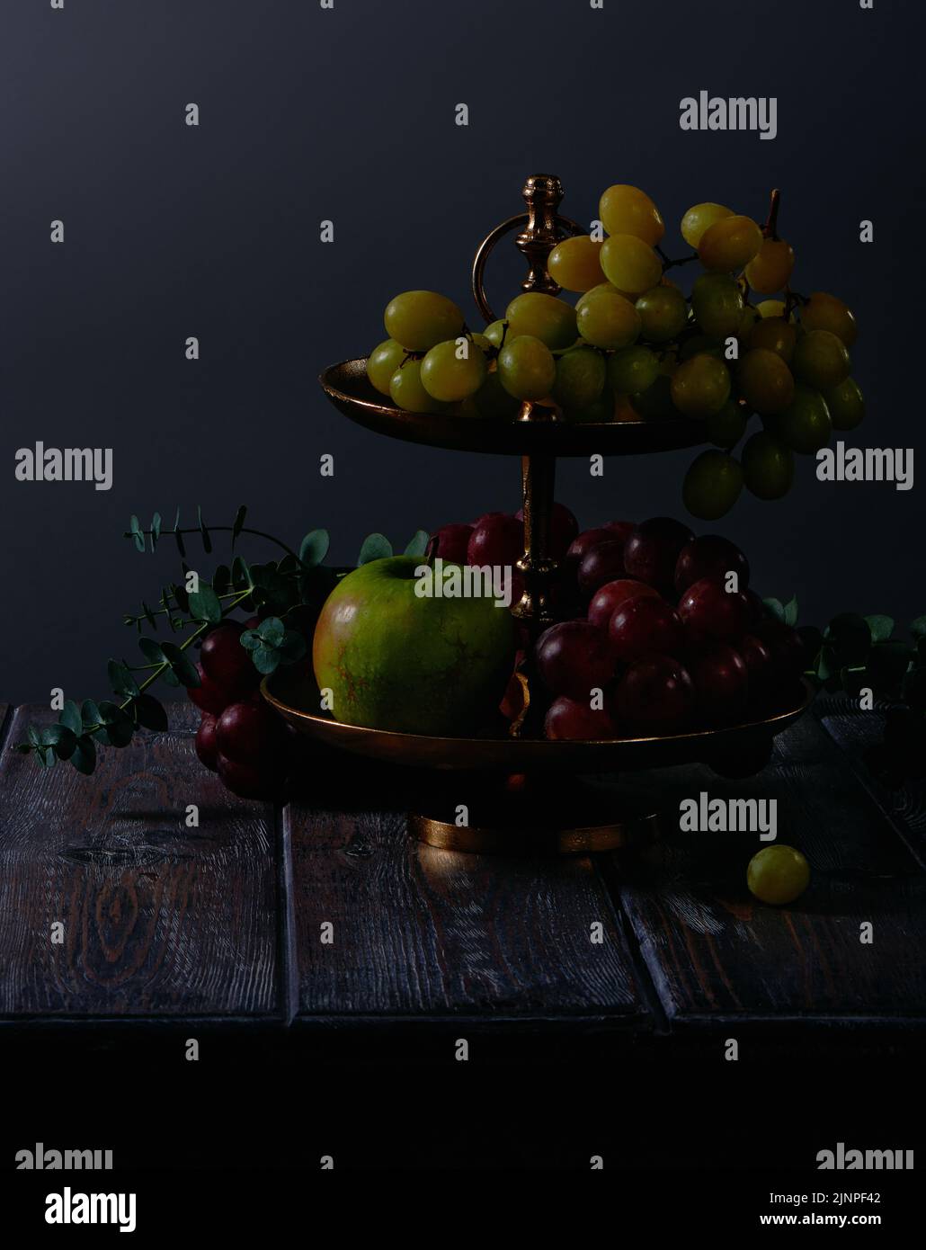 Fruit still life with grapes, apples and eucalyptus in antique copper dessert stand on dark wooden table background Stock Photo