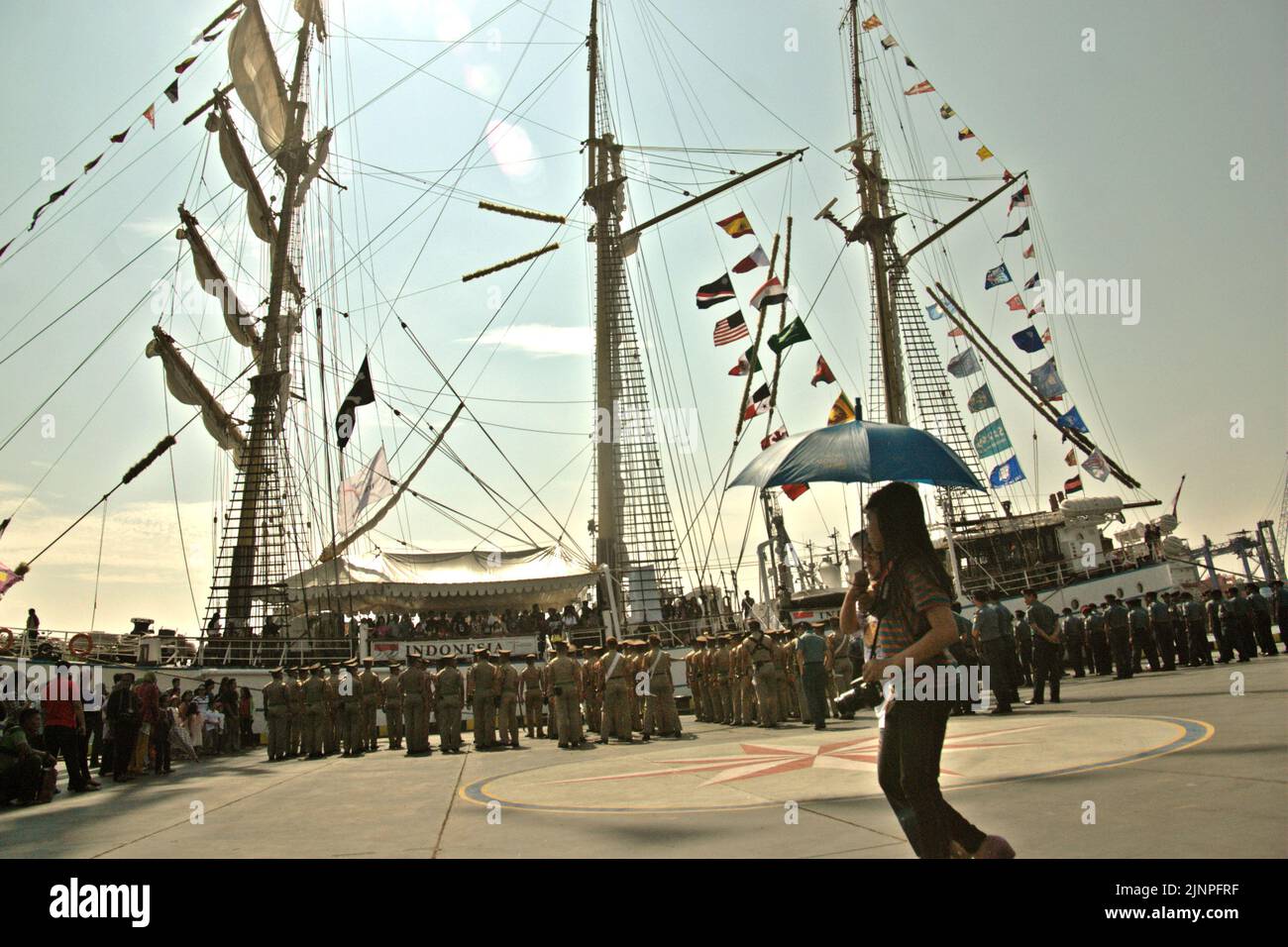 Young women carrying an umbrella and a camera, as they are walking on the dock, in a background of KRI Dewaruci (Dewa Ruci), an Indonesian tall ship, that is being opened for public visitors at Kolinlamil harbour (Navy harbour) in Tanjung Priok, North Jakarta, Jakarta, Indonesia. Stock Photo