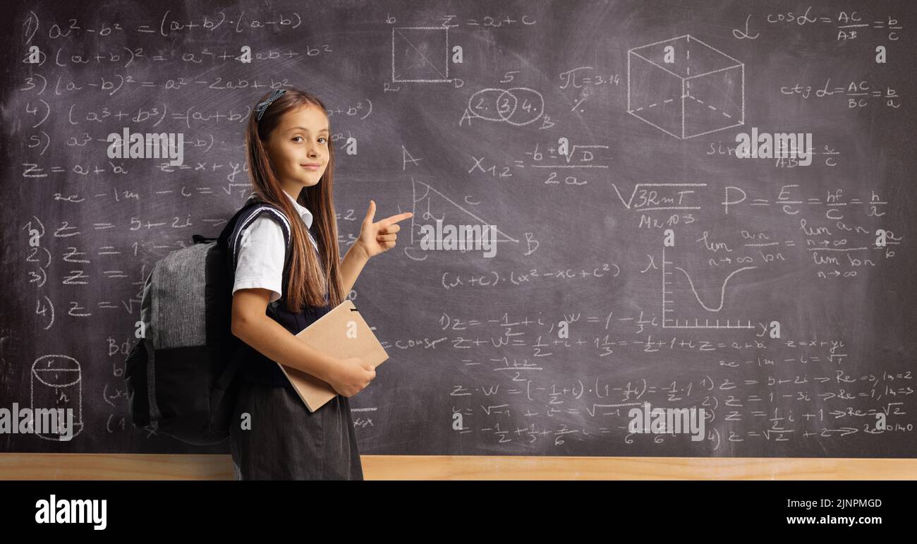 Female pupil in a school uniform with a backpack and book pointing at a blackboard with numbers and math formulas Stock Photo