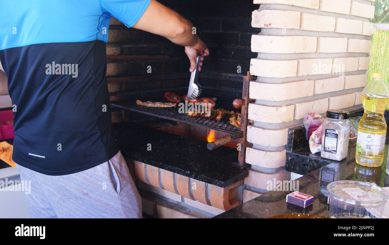 Young man roasting meat and sausage on barbecue grill with knife and fork with teeth, Brazil, South America, zoom photo Stock Photo