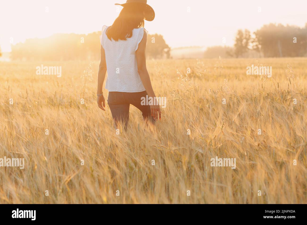 Woman farmer in cowboy hat walking with hands on ears at agricultural field on sunset. Stock Photo