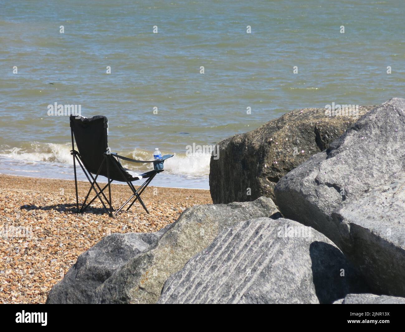 The perfect spot: a single folding camping chair on the sand facing the gently lapping waves of the sea with nearby rocks; the beach at Felixstowe. Stock Photo