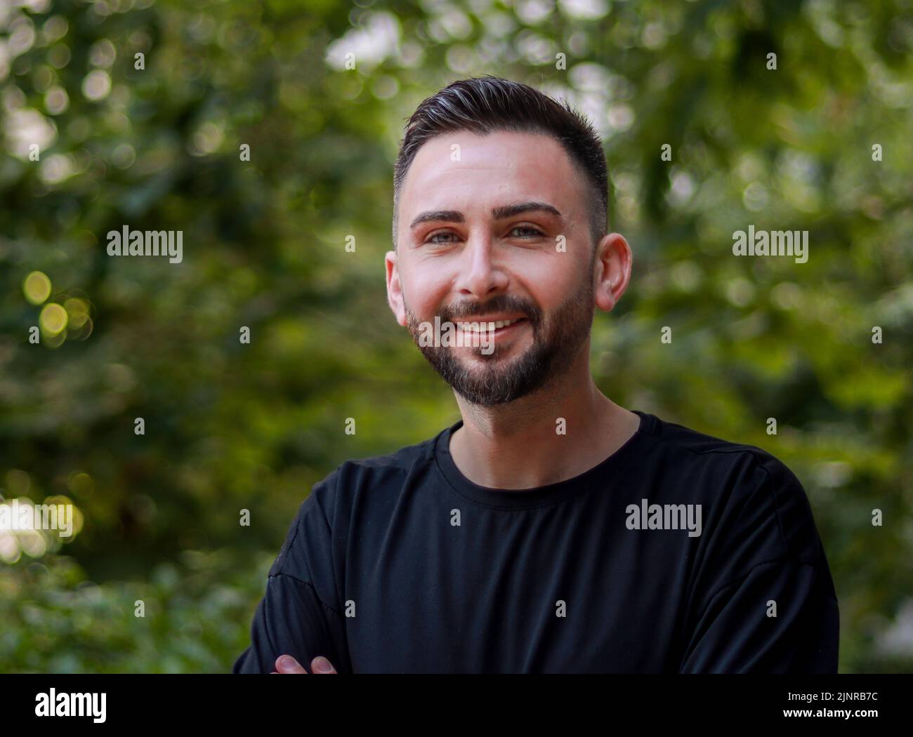 Portrait of young handsome man with beard. Young man in black shirt smiling looking at camera. Outdoor nature background Stock Photo