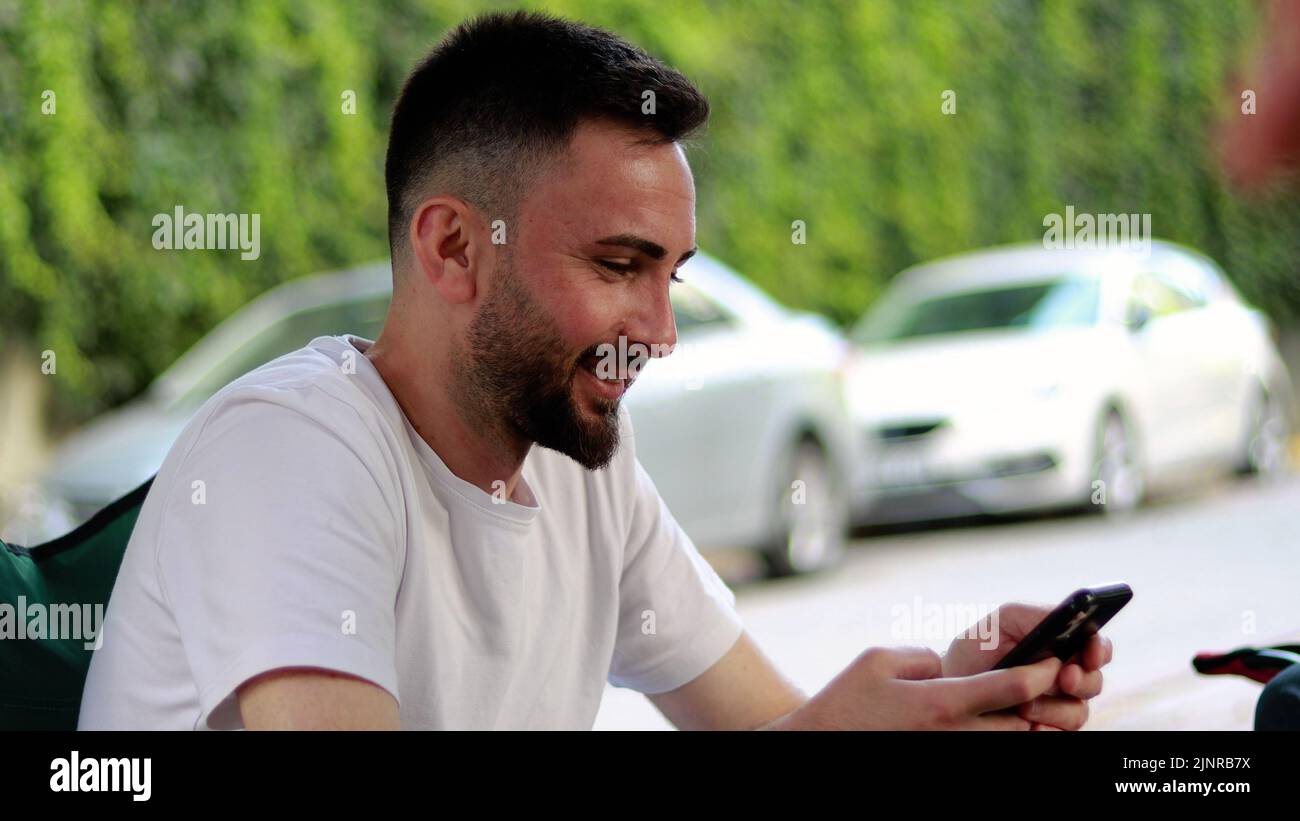 Bearded and handsome young man using mobile phone outdoors. Happy smiling man in white t-shirt looking at mobile phone screen and reading messages Stock Photo