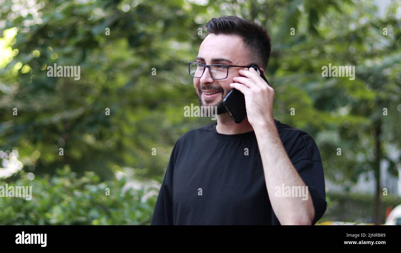 Portrait of young handsome man talking on the phone with beard and glasses. Smiling young man in black shirt. Outdoor nature background Stock Photo