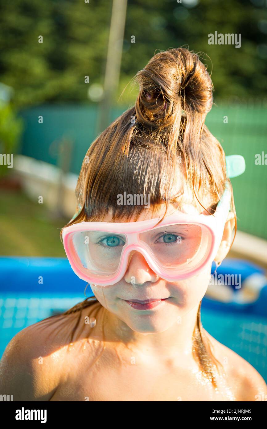 Caucasian child girl with hair bun and funny expression wearing swimming goggles in home pool. Outdoor portrait. Vertical. Stock Photo