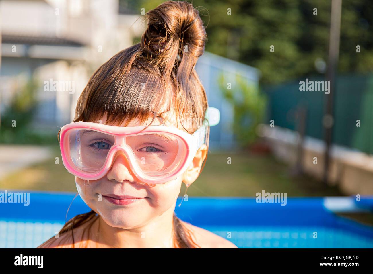 Caucasian child girl with hair bun and funny expression wearing swimming goggles in home pool. Outdoor portrait. Stock Photo