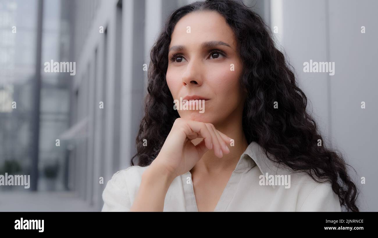 Close up pensive thoughtful puzzled Caucasian Hispanic thoughtfully girl woman with curly hair standing outdoors sad worry dreaming planning Stock Photo