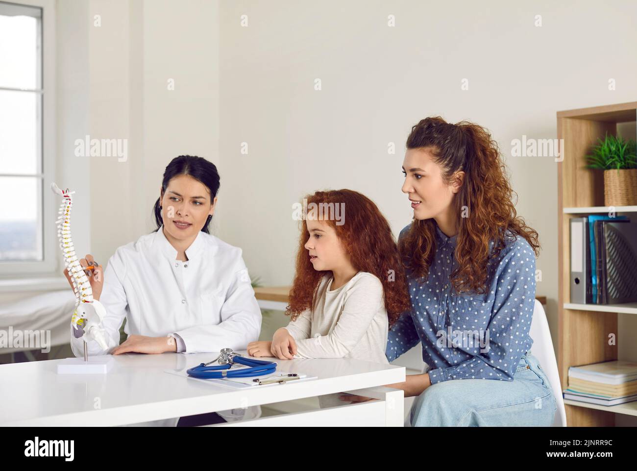 Doctor at orthopedic clinic talking to mother and child and showing anatomical spine model Stock Photo