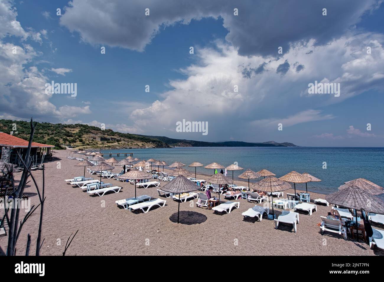 Parasols and lounge chairs on the beach with turquoise colored water of Assos on Aegean Sea. People tanning in summer day. Stock Photo