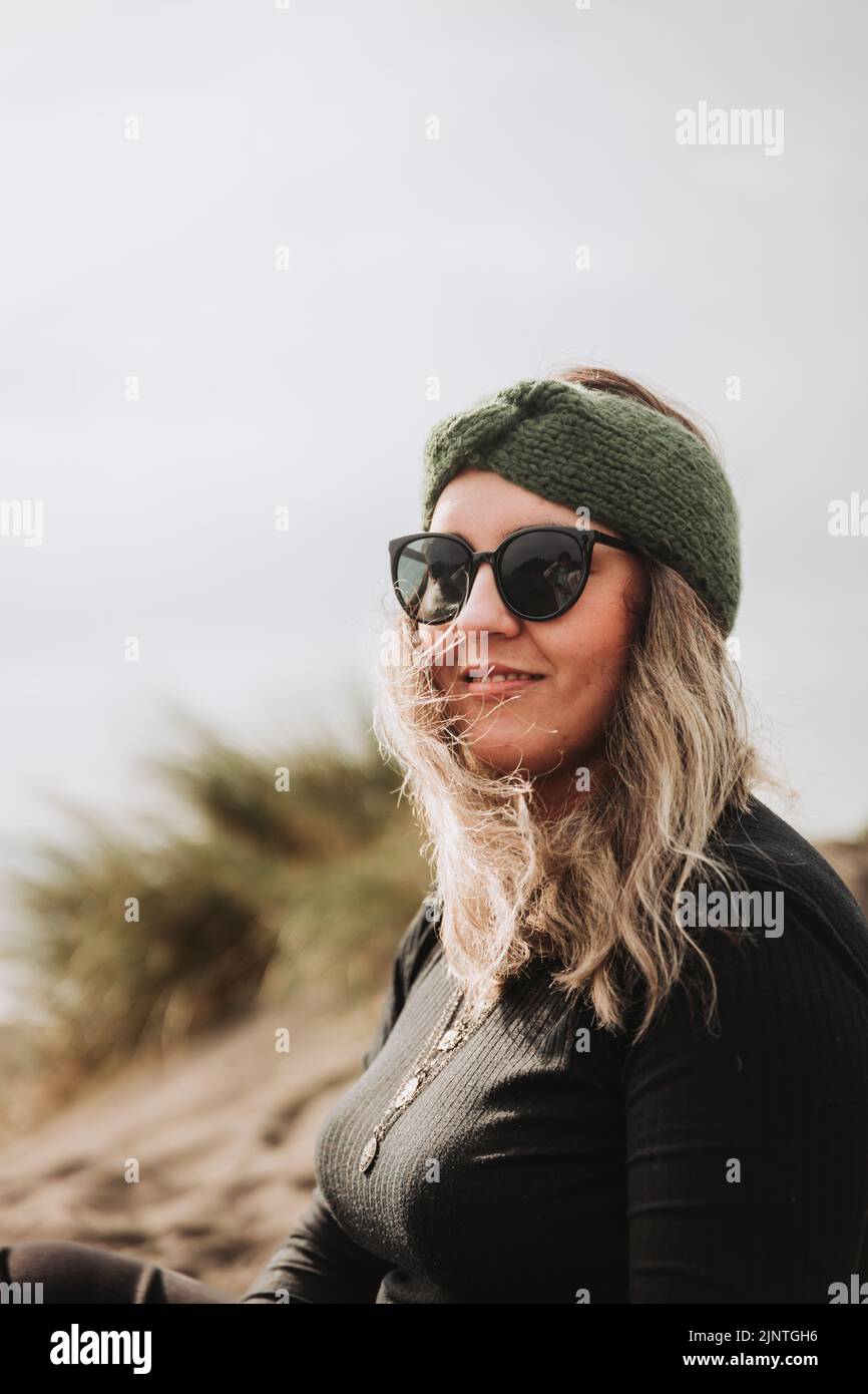 Smiling blonde woman dressed in black, wearing a headband, sunglasses and sitting on the beach. Overcoming depression Stock Photo
