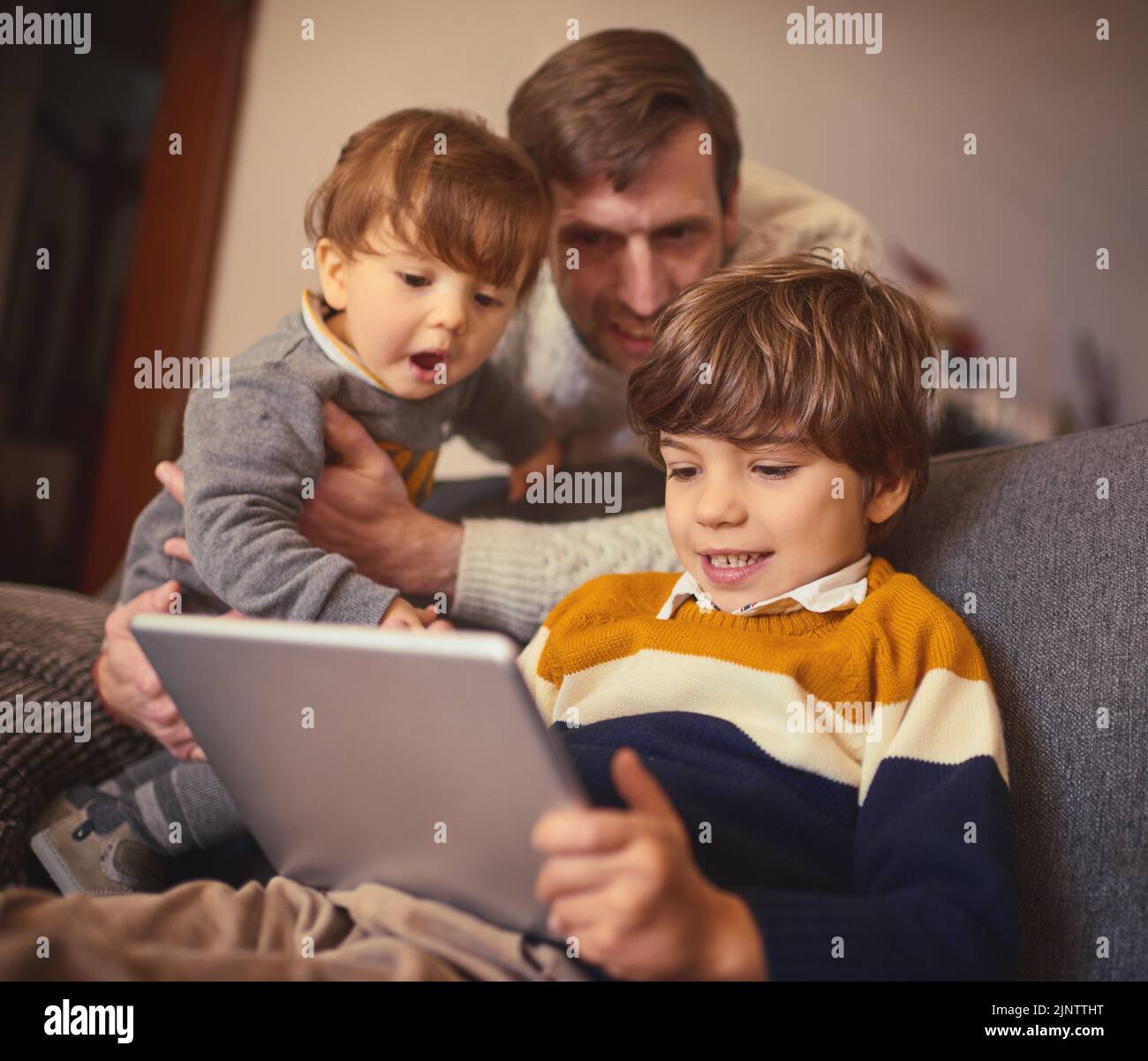 Lets see what your brothers doing. an adorable little boy using a tablet on the sofa at home while his father and younger brother look on. Stock Photo
