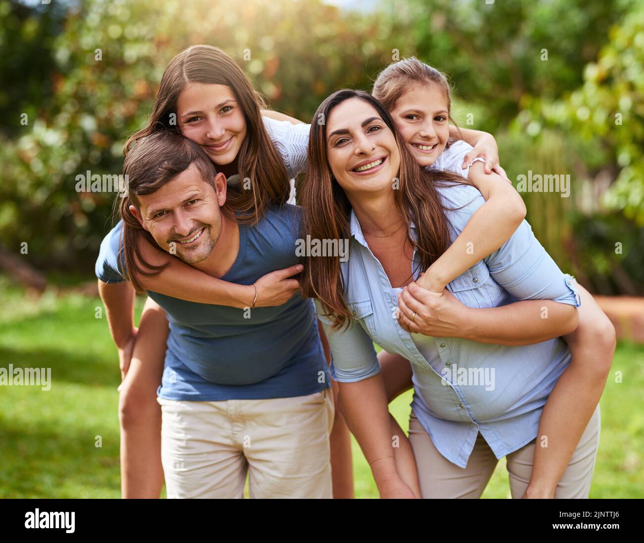 Lets take a ride. Portrait of a cheerful young mother and father giving their daughters a piggyback ride outside in a park during the day. Stock Photo