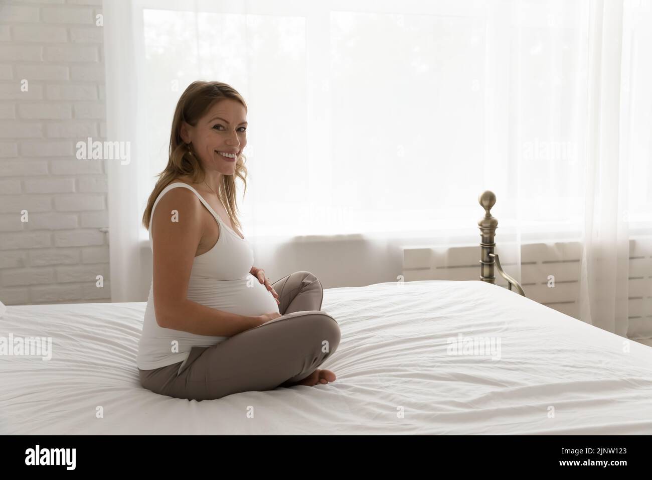 Attractive pregnant woman sits on bed smile looks at camera Stock Photo
