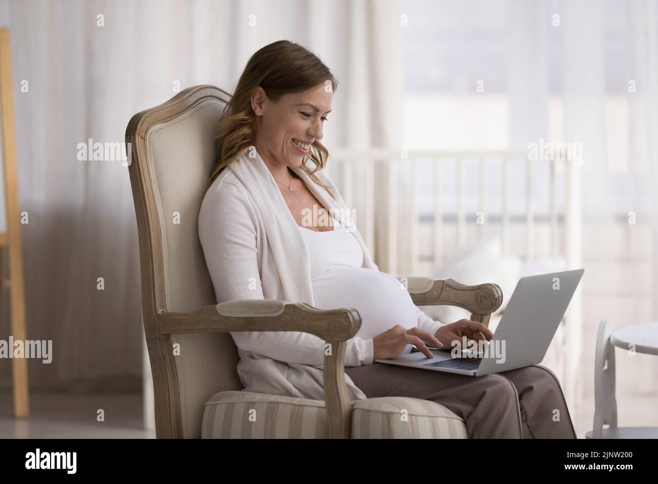 Young pregnant smiling woman relax on armchair with laptop Stock Photo