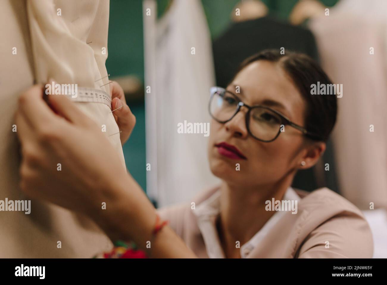 Young woman dressmaker taking measurements of dress on mannequin in her studio. Asian fashion designer adjusting dress on a model. Stock Photo