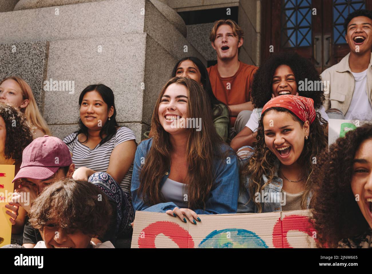 Group of youth activists smiling happily while sitting outside a building with posters and banners. Multicultural young people protesting against glob Stock Photo