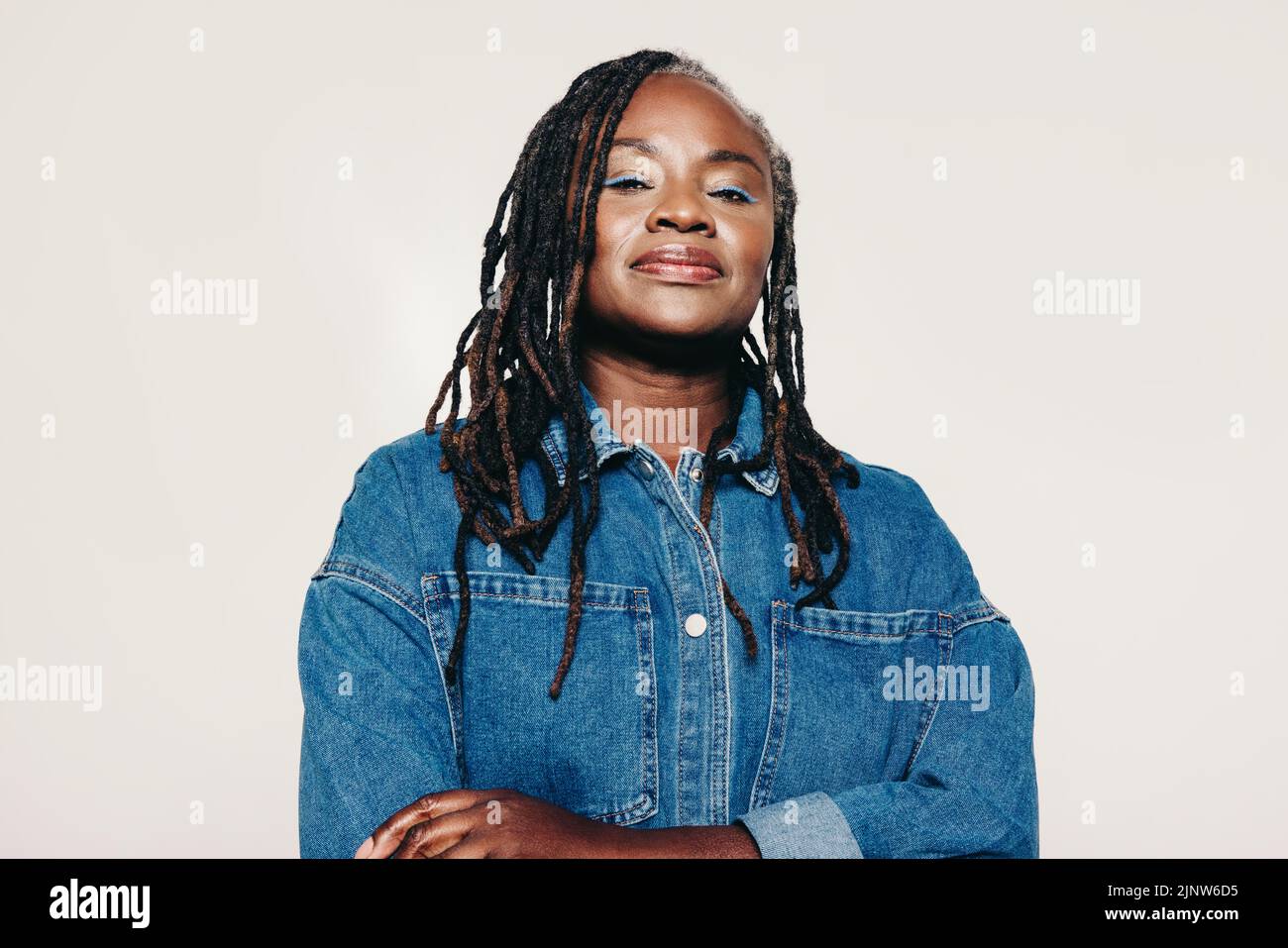 Confident mature woman looking at the camera while standing against a studio background with her arms crossed. Fashionable woman with dreadlocks weari Stock Photo