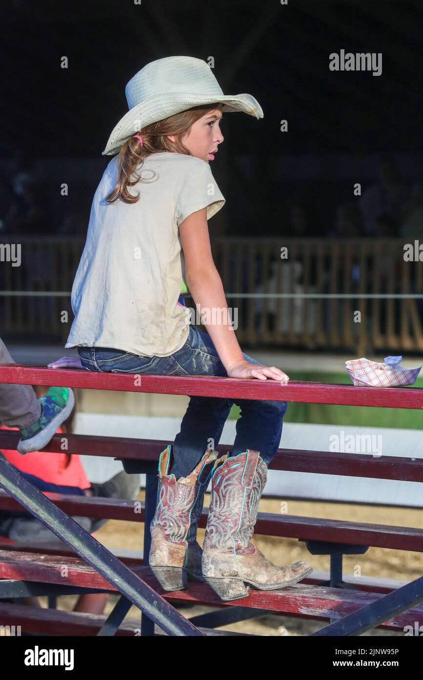 A girl wearing a cowboy hat and cowboy boots watches the mini horse