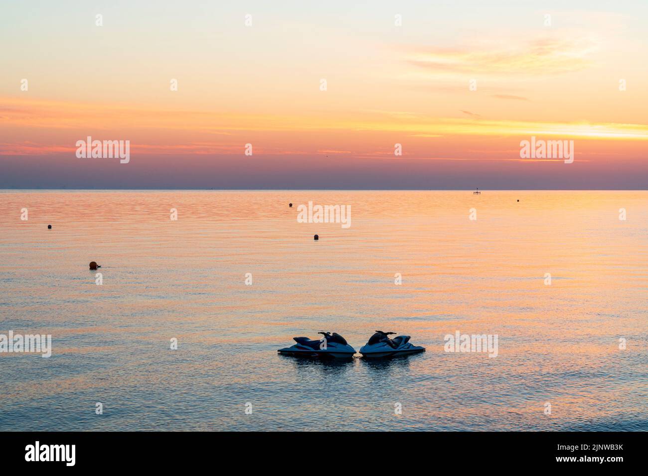 Dawn sky over the North Sea seen from Herne Bay on the English Kent coast. Orange sky with the colour reflecting in the calm water, and in the foreground a pair of jet skis moored in the sea and the main beach. Stock Photo