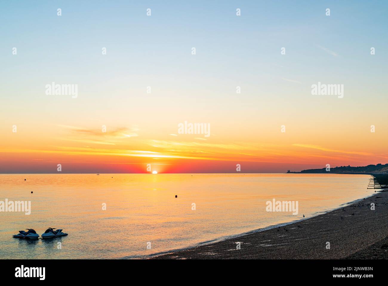 Sunrise over the North Sea seen from Herne Bay on the English Kent coast. Sun rising into an orange sky and in the foreground a pair of jetskis moored in the sea and the main beach. Stock Photo