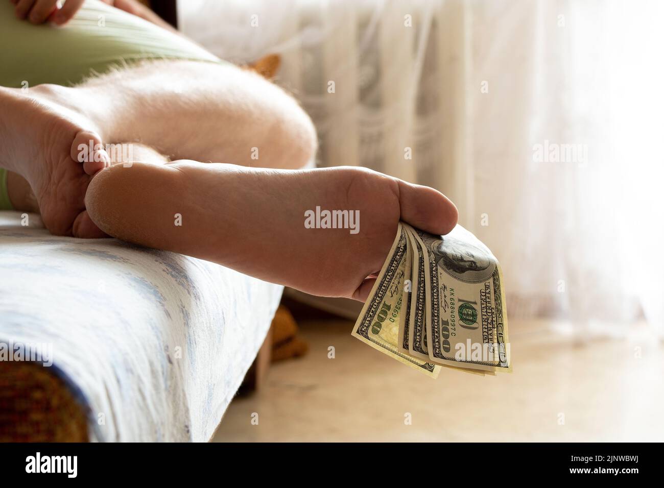 Man's foot holding one hundred dollars with toes lying on the bed at