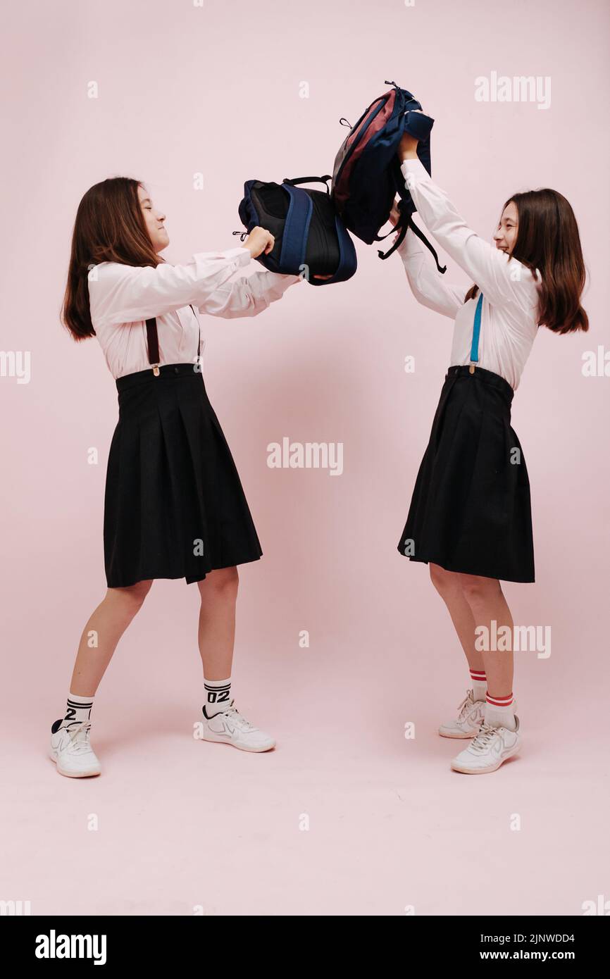 Fighting playful schoolgirl twins bashing each with their backpacks for fun. Wearing school uniforms with knee high skirts and suspenders of different Stock Photo