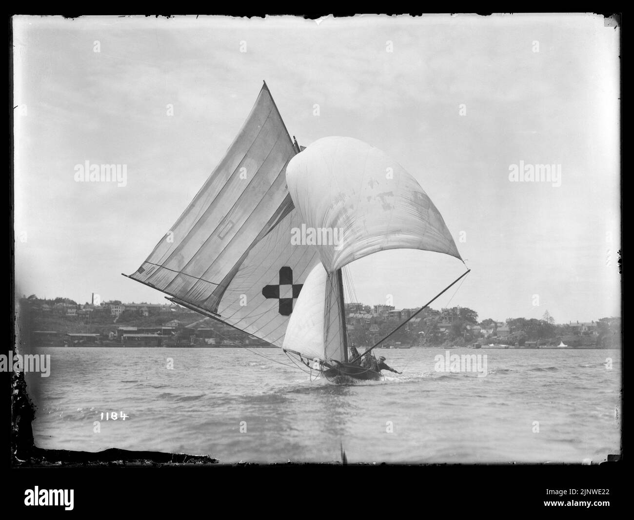 12 footer COMMONWEALTH on Sydney Harbour *** Local Caption *** Glass plate negative. Hall, William. Untitled (12 footer, has a cross with a centre square on main sail). Sydney harbour, New South Wales, Australia. Glass plate negative. Viewed from infront, flying spinnaker. Buildings visible on the land in the background. Inscription '1184' l.l. Stannard collection. Stock Photo