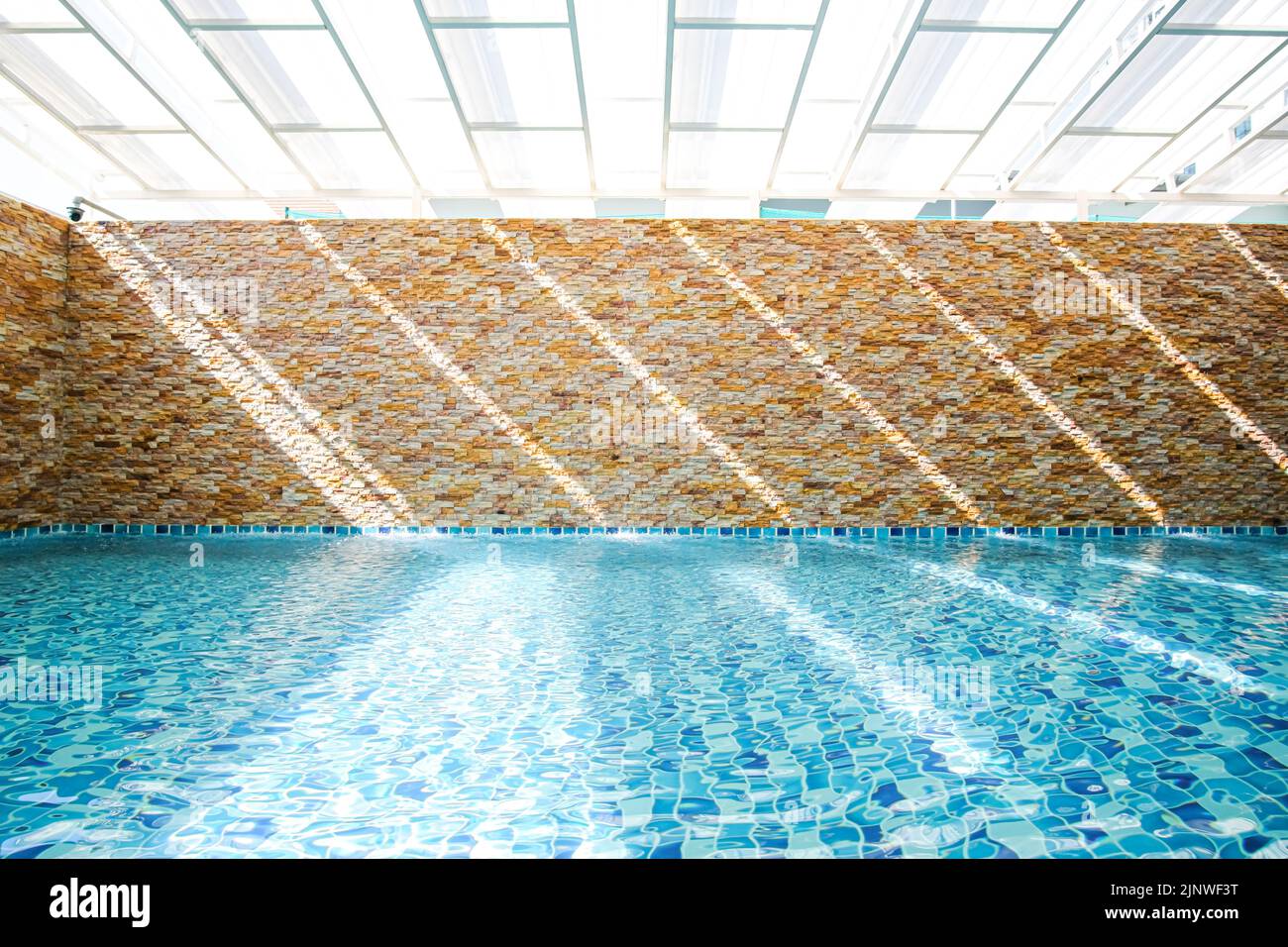 In an empty turquoise swimming pool, sunlight shines through a translucent roof on the surface of the water and a stone wall in the backdrop. Stock Photo