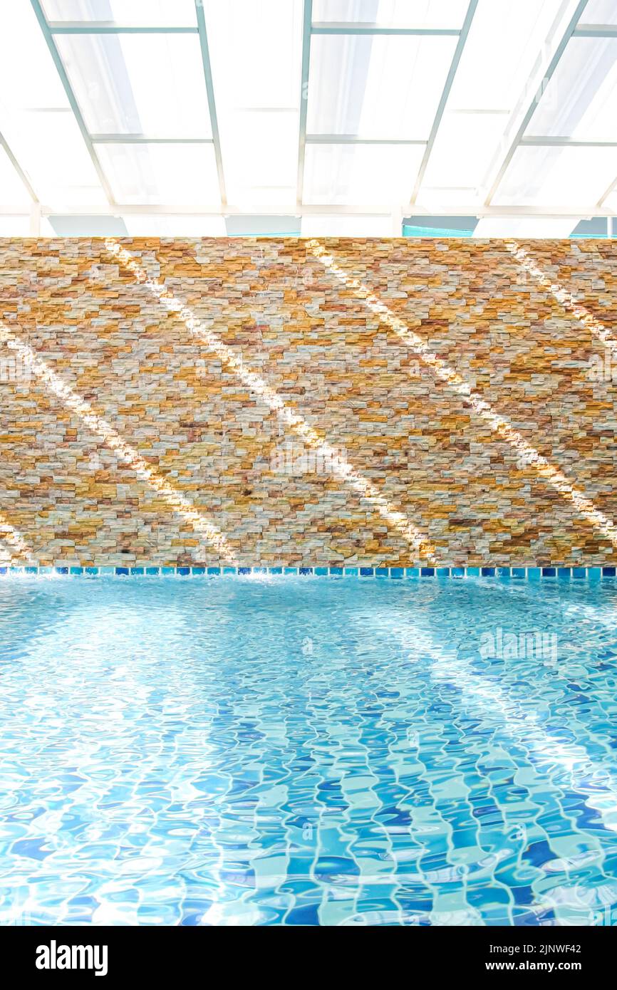 In an empty turquoise swimming pool, sunlight shines through a translucent roof on the surface of the water and a stone wall in the backdrop. Stock Photo