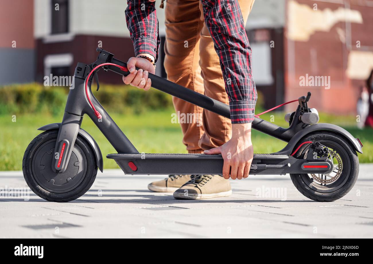 Folded electric scooter on concrete pavement, young man behind holding bent front bar, closeup detail Stock Photo